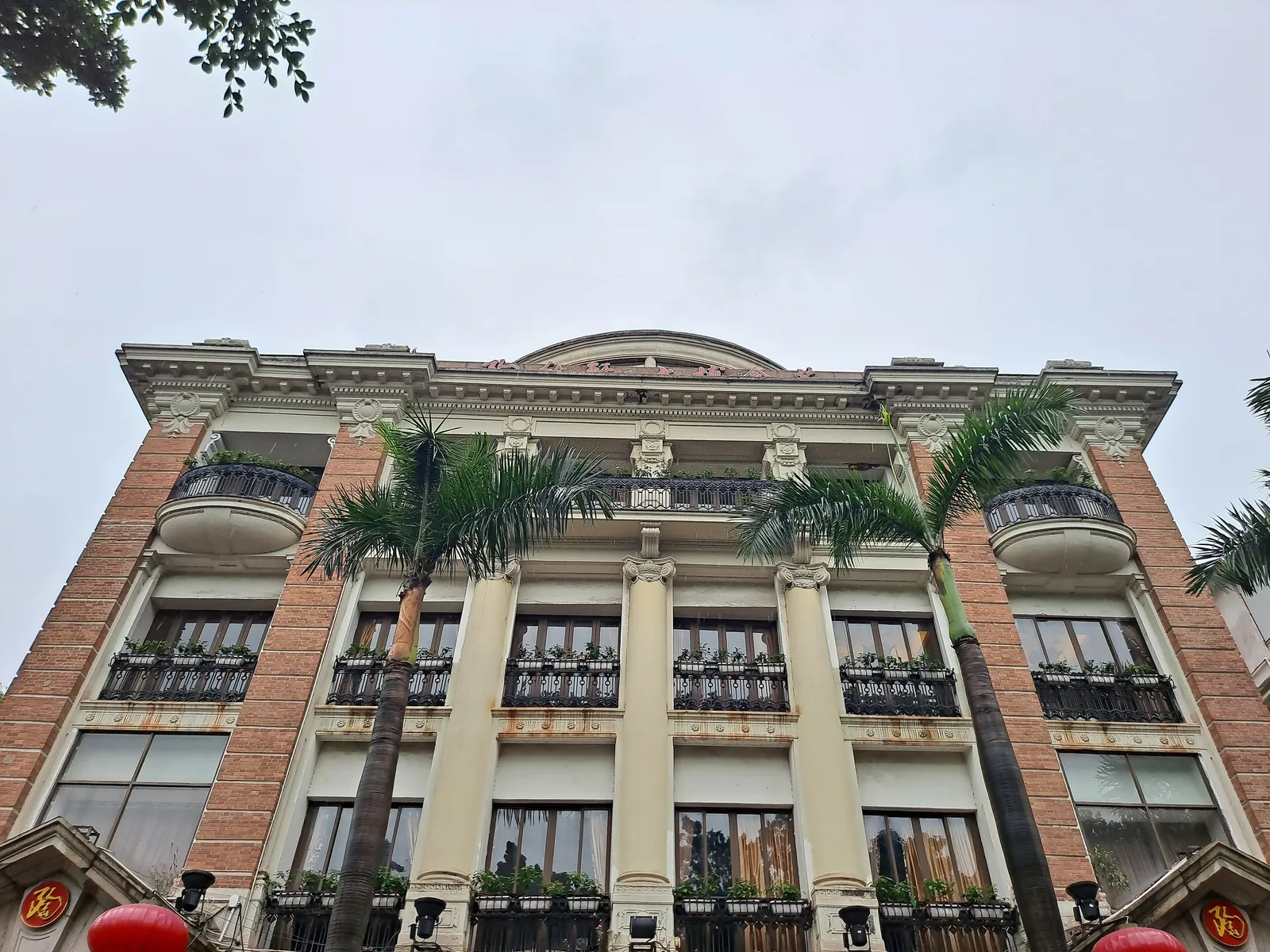 A pale building of four storeys with brick corner piers, rounded corner balconies and palm trees in front.