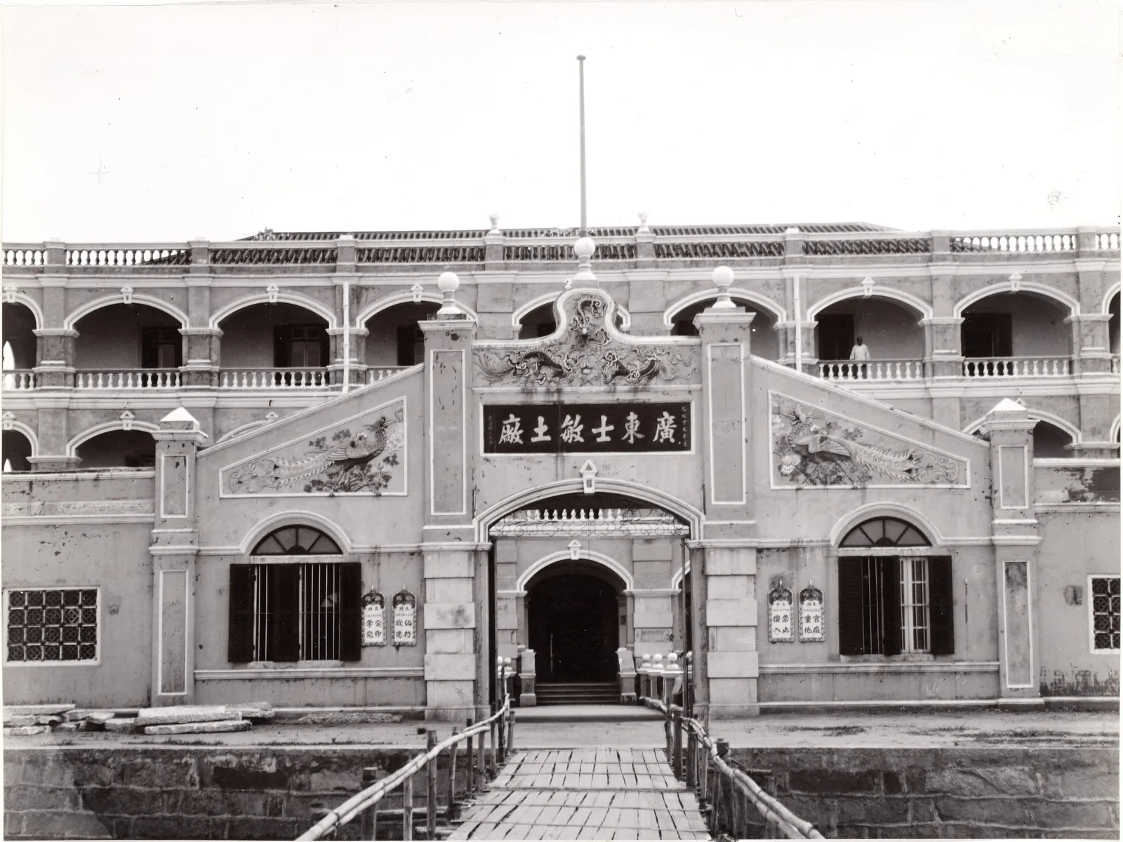 A rendered gatehouse with a carved signboard and a gangway to its arched door, with two arcaded buildings behind.