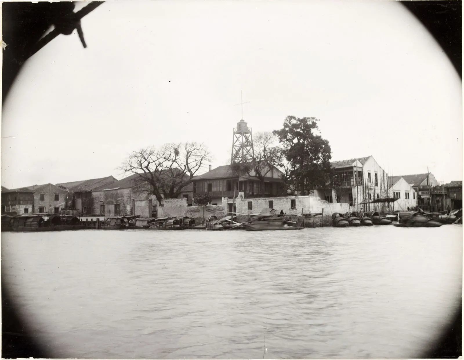 Low buildings along a river bank, seen from the water through a round vignette.