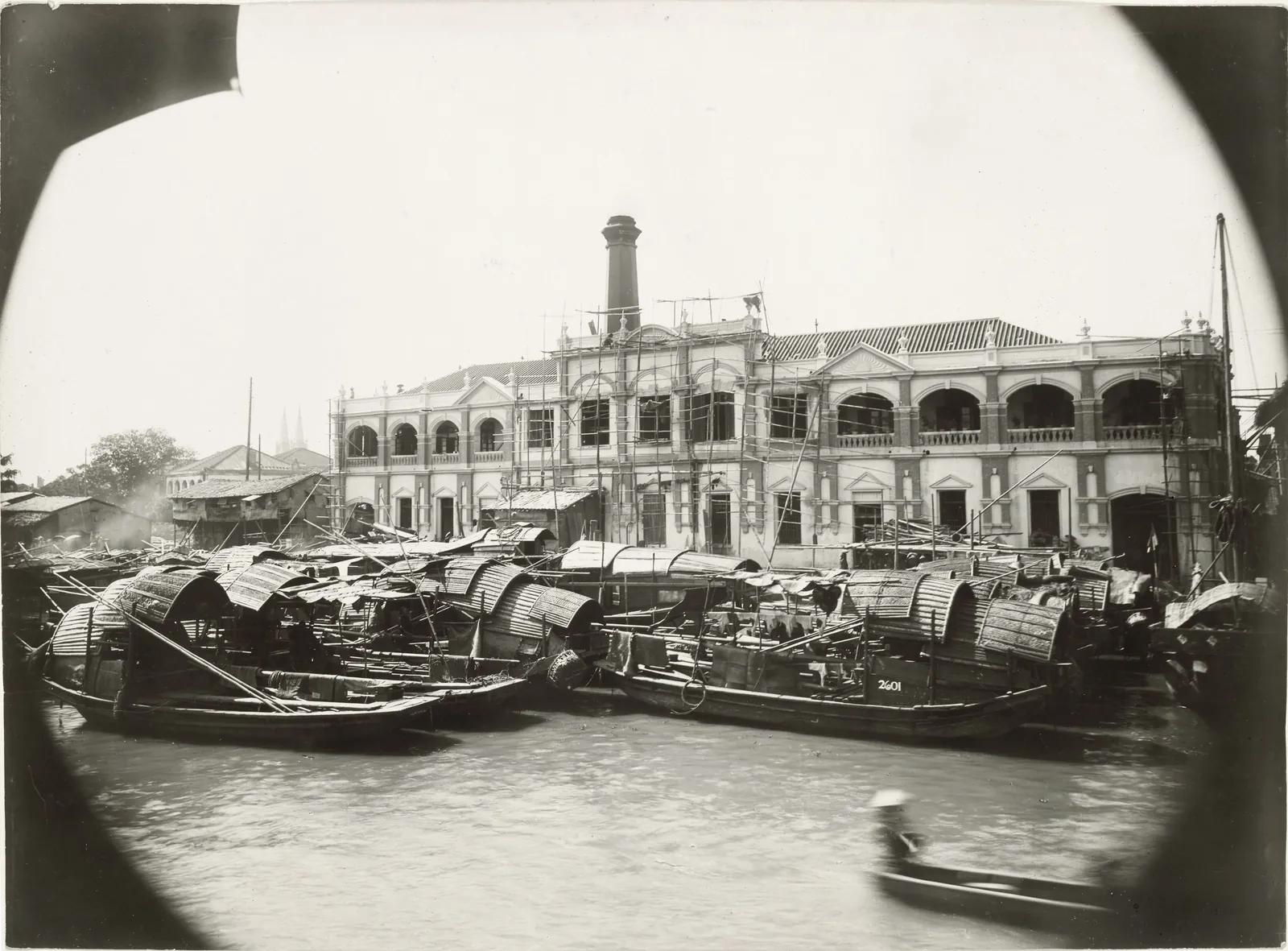 A two-storey arcaded building with a tall chimney behind, seen across the river over a crowd of moored sampans.